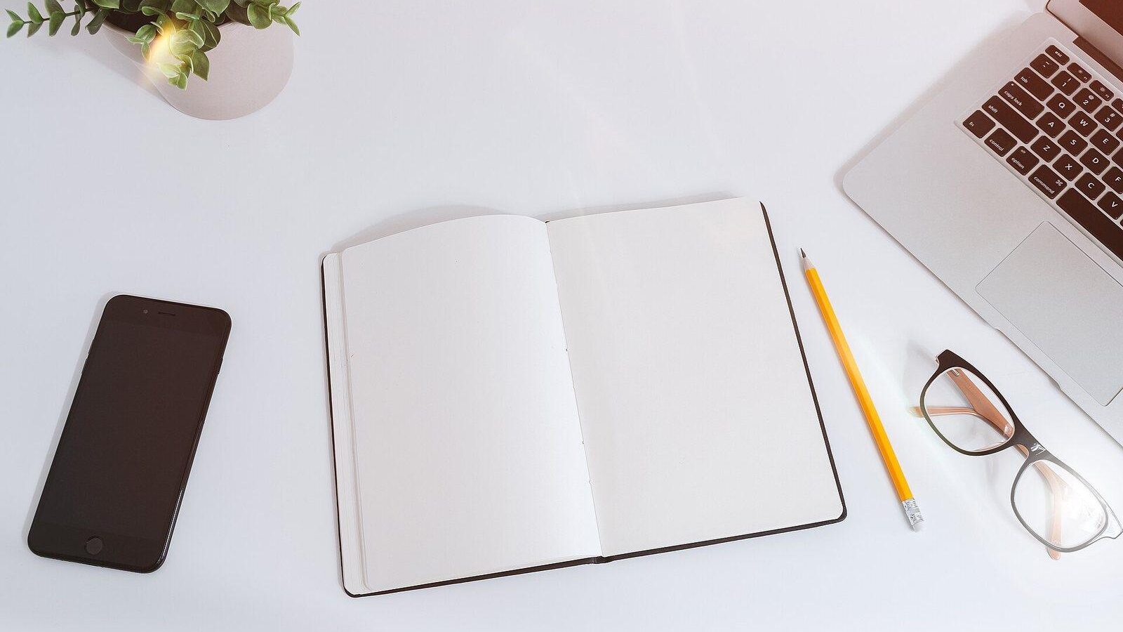 Minimal white desk with a smartphone, open notebook, pencil, glasses and laptop