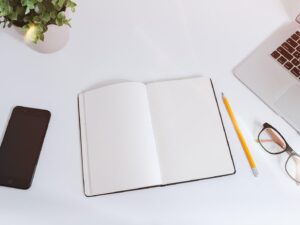 Minimal white desk with a smartphone, open notebook, pencil, glasses and laptop