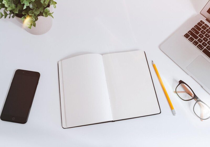 Minimal white desk with a smartphone, open notebook, pencil, glasses and laptop