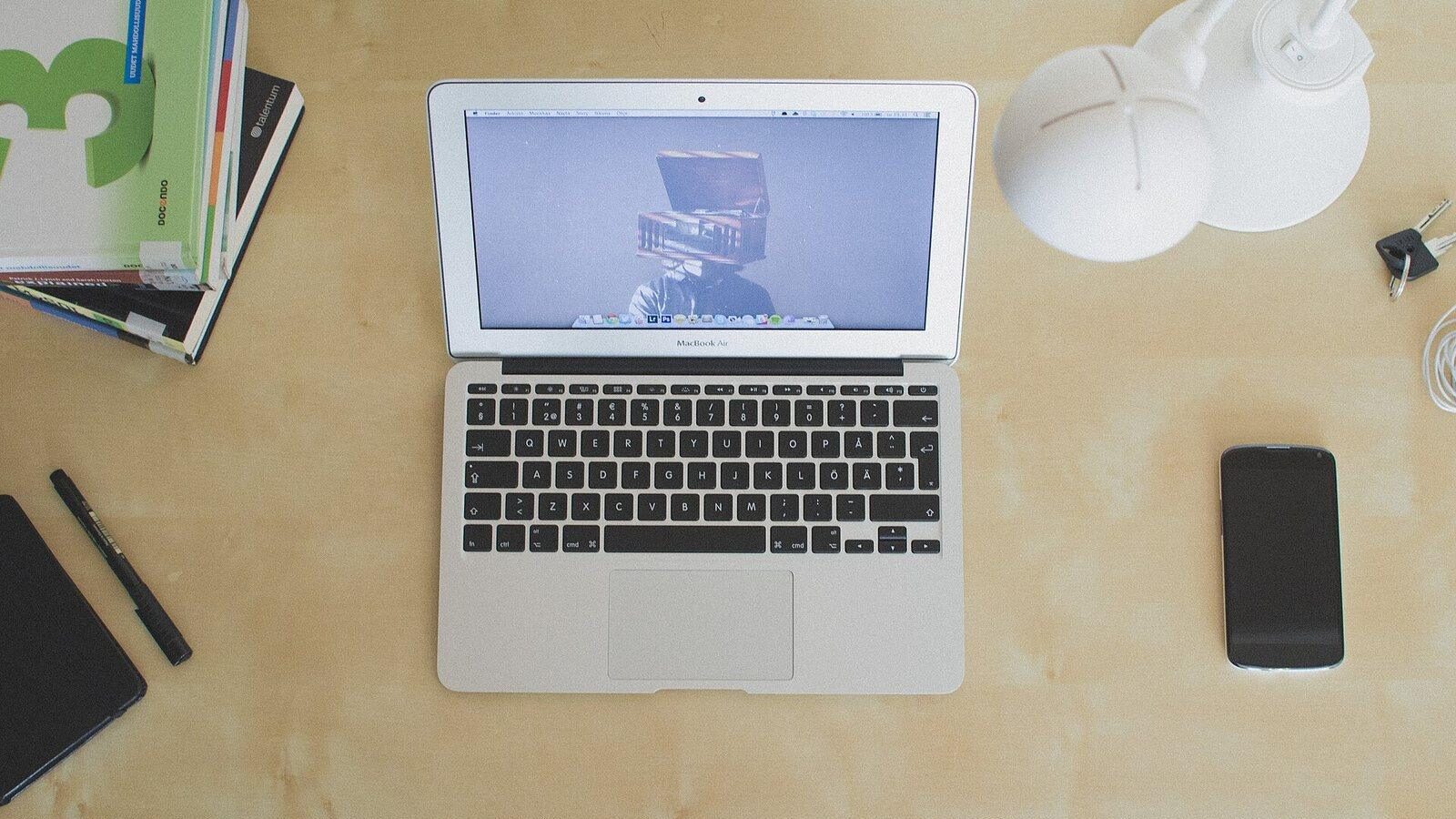 Top-down view of an open laptop on a light wooden desk with a phone and notebooks