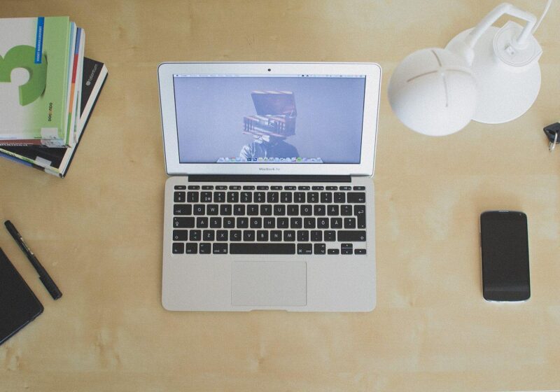 Top-down view of an open laptop on a light wooden desk with a phone and notebooks