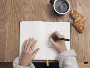 Person writing meeting notes in an open notebook next to a cup of coffee