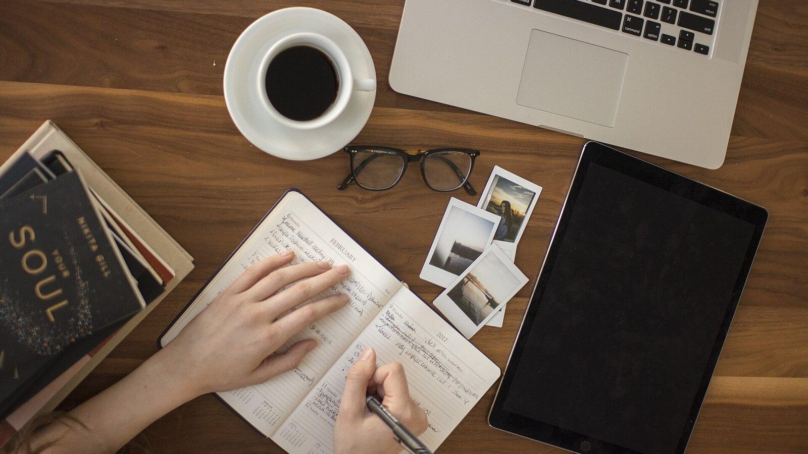Hands writing in a journal beside a coffee cup, laptop and tablet on a wooden desk