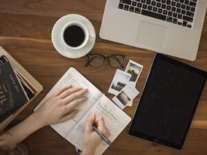 Hands writing in a journal beside a coffee cup, laptop and tablet on a wooden desk