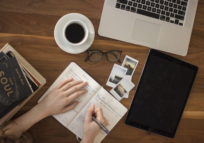 Hands writing in a journal beside a coffee cup, laptop and tablet on a wooden desk